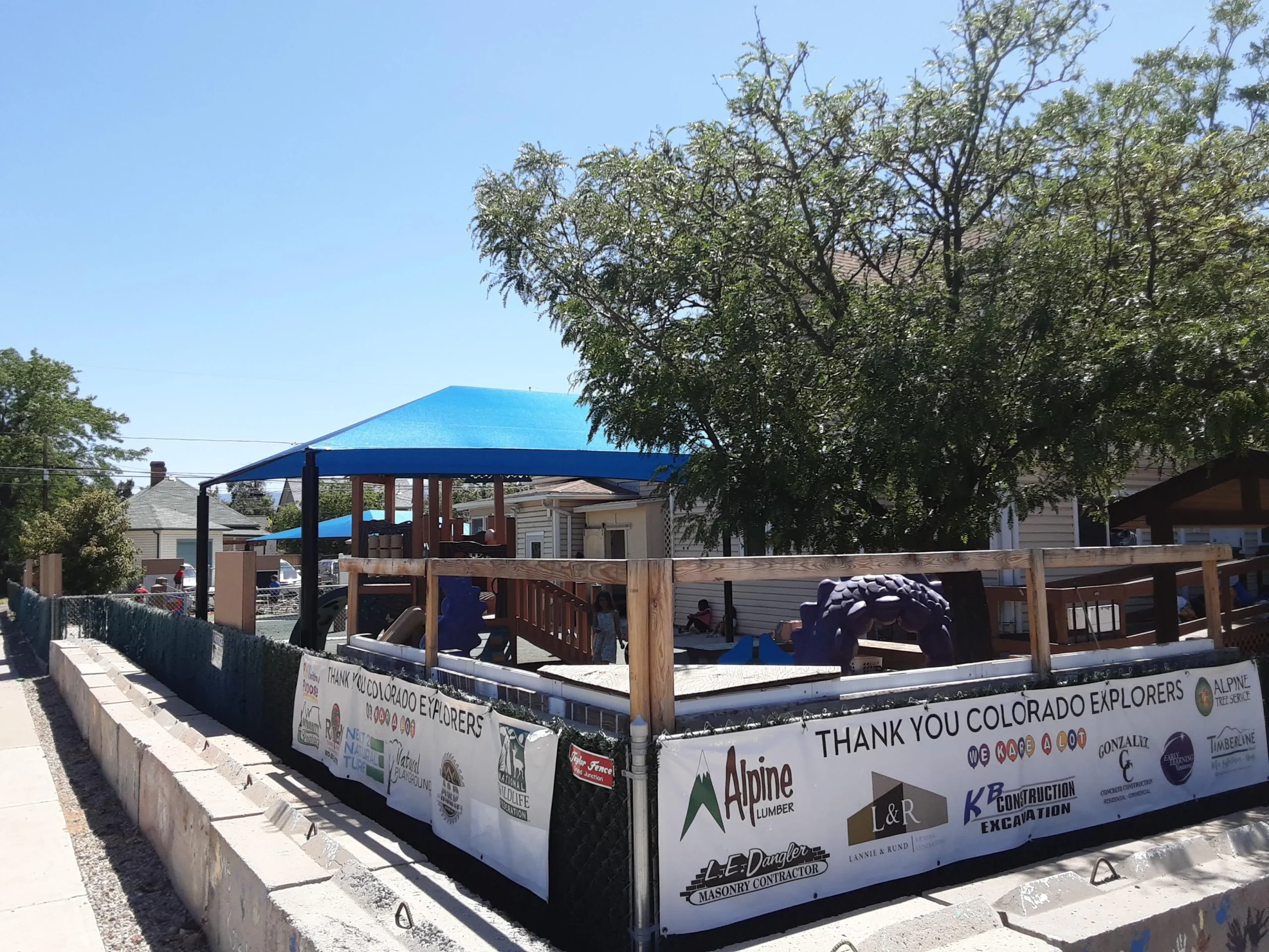 An outdoor fenced playground with a blue shade canopy and sponsorship banners along the perimeter