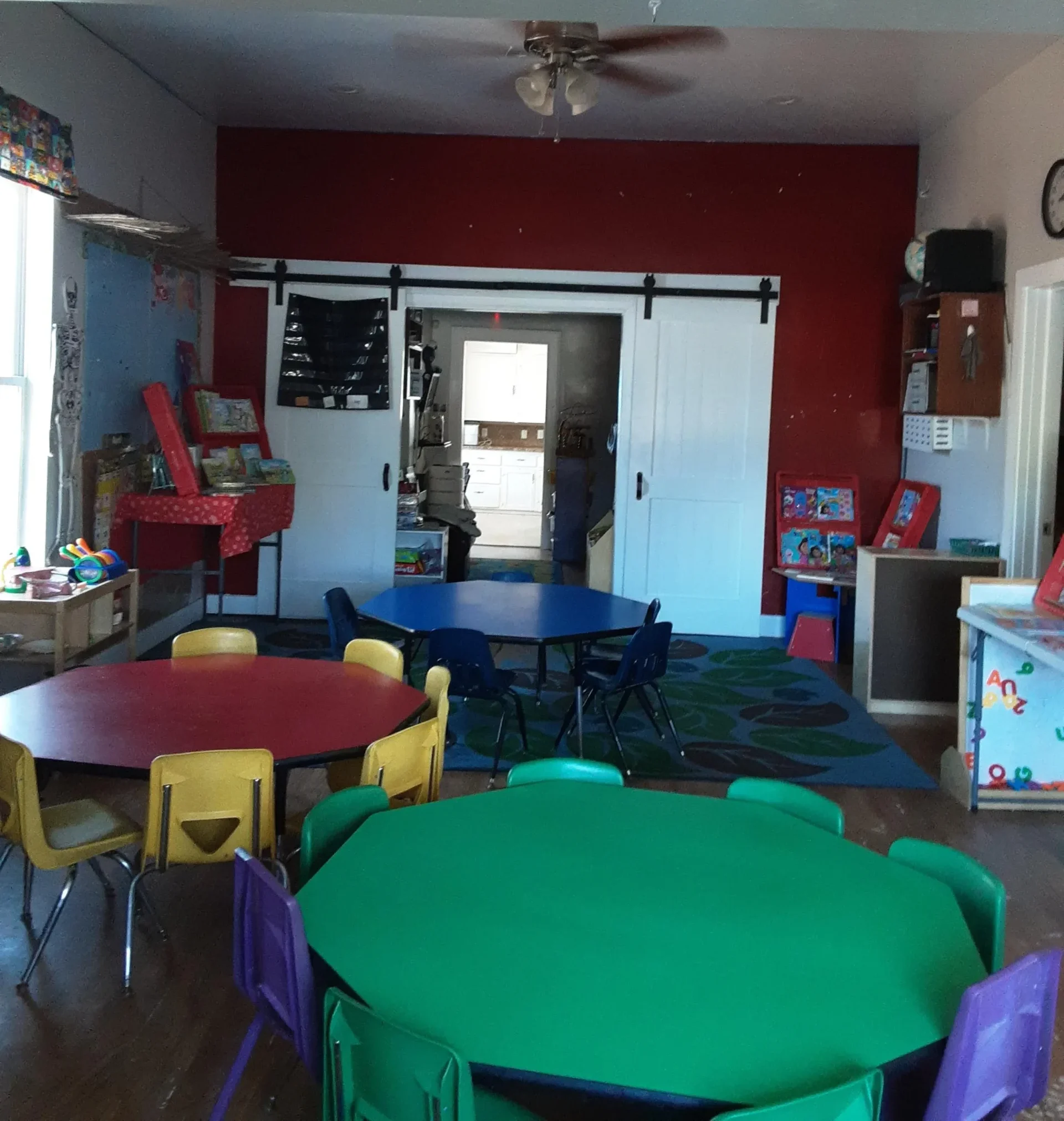 A colorful early childhood classroom with low green, red, and blue activity tables and small chairs
